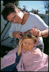 It is my first morning home after being in isolation and two weeks since my first chemo infusion. Each morning I wake up and gently tug at my hair. So far it has stayed put but this morning it gives way and comes out in clumps. For the last week my scalp has been tender to the touch and very itchy. I study my pillow and find it is covered with hair. I swear, which doesn’t help but makes me feel better. Then I pray for the strength to get through the months to follow. I make my way downstairs, running my hand through my hair as I go. More comes away in my fingers so I swear again. “Fucking cancer!” I make a bowl of cereal and take it outside to eat. There is a soft breeze and I watch more hair fall into my breakfast then contemplate the dark strands floating on my cup of tea. After a minute I tip my drink and cereal onto the garden and move inside. I phone Helen and ask what she is doing. She lives ten houses up the road. I tell her I need her help to chop my hair off. She doesn’t say much, just arrives soon after. I comment I should phone Tim so he can take photos. He joins us moments later. The three of us sit in the garden. Helen drapes a towel around my shoulders. I’m not allowed to see the progress until the job is finished. I don’t want to look at myself anyway. I’m scared of what I might see. A pile of hair lies on the grass like the remains of a catfight. On the far side of the garden wall, cars pass in noisy succession. Life goes on. On this side Tim’s camera clicks repeatedly. The three of us laugh and make jokes. My own laughs drown out the anxious thudding of my heart. My own Mum whispers from somewhere in my ear, ‘This too will pass,’ and I take heart knowing she is here in my garden with me. I smile to myself thinking she would have heard me swearing this morning. She always said I was lazy with my adjectives. The chunky sewing scissors Helen used have left ridges and patterns in my remaining hair. It is cropped as close to my scalp as she can get it. Her verdict is, “It’s quite funky,” but I’ll be the judge of that. I want to be alone to take the change in. Helen says goodbye and Tim follows me upstairs to the mirror. He quietly snaps a few photos then leaves me to my thoughts. My hair is very dark. That is a surprise. My eyes look haunted and stare out from my swollen pale face. When I am through the initial shock of it I begin to think I am lucky. My head is small and nicely shaped. No fat wrinkles at the back of my neck, just a pale strawberry birthmark. My ears lie flat against my head and, for now at least, I still have my eyebrows and eyelashes. Some makeup will do wonders. Yes, I can live with this. To prove it I set myself a challenge – to go out this very afternoon to Merivale Mall with no hat on. I think if I can do that then everything from now on will be all right. Two weeks ago I wouldn’t have remotely entertained he thought. I ring Helen and tell her of my plan. She laughs and tells me she can meet me for a stiff drink afterwards if I need one. Within the month I have no hair at all. My scalp is shiny white. My eyebrows are thinning and my eyes are constantly itchy and watering. I have very little to hang my mascara on but I persevere despite it all. I will not paint eyebrows on though. That is one thing I will not do. I joke with Tim that I haven’t just lost the hair on my head, the hair on my lower body has fallen out too. Having no pubic hair is pretty strange, God, even the fine hairs on my bum have fallen out. – Ib